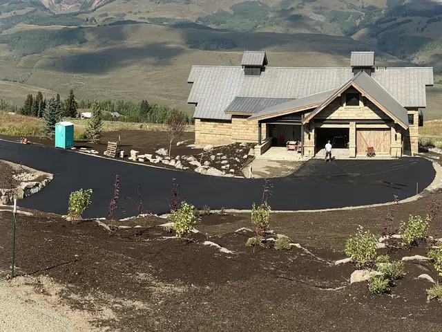 Fresh asphalt driveway curving up to a stone and timber mountain home, with the paving crew working at its far end