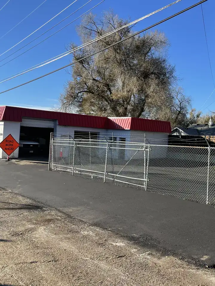 New asphalt lot along a fenced shop building under a big cottonwood