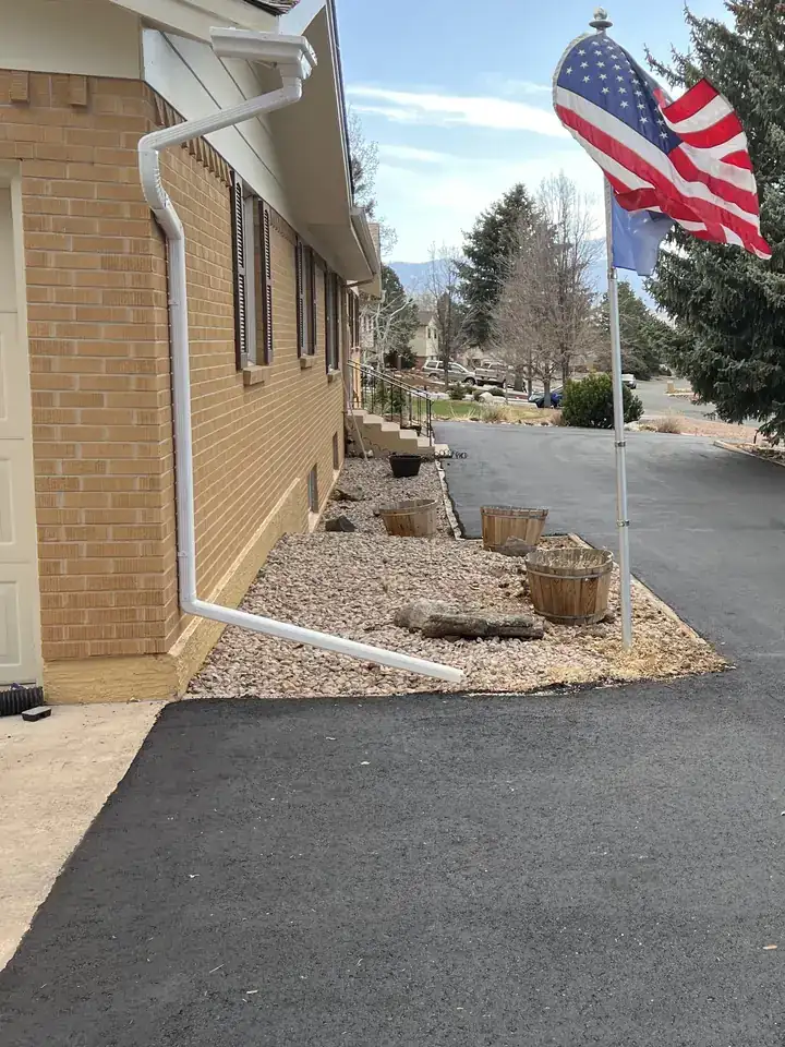 New black asphalt driveway beside a brick rambler with rock landscaping and blue spruce